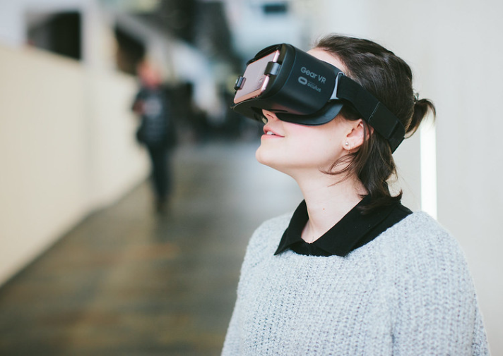 Eine weiße Frau mit braunen Haaren und hellem Pullover trägt eine VR-Brille und schaut leicht nach oben. 