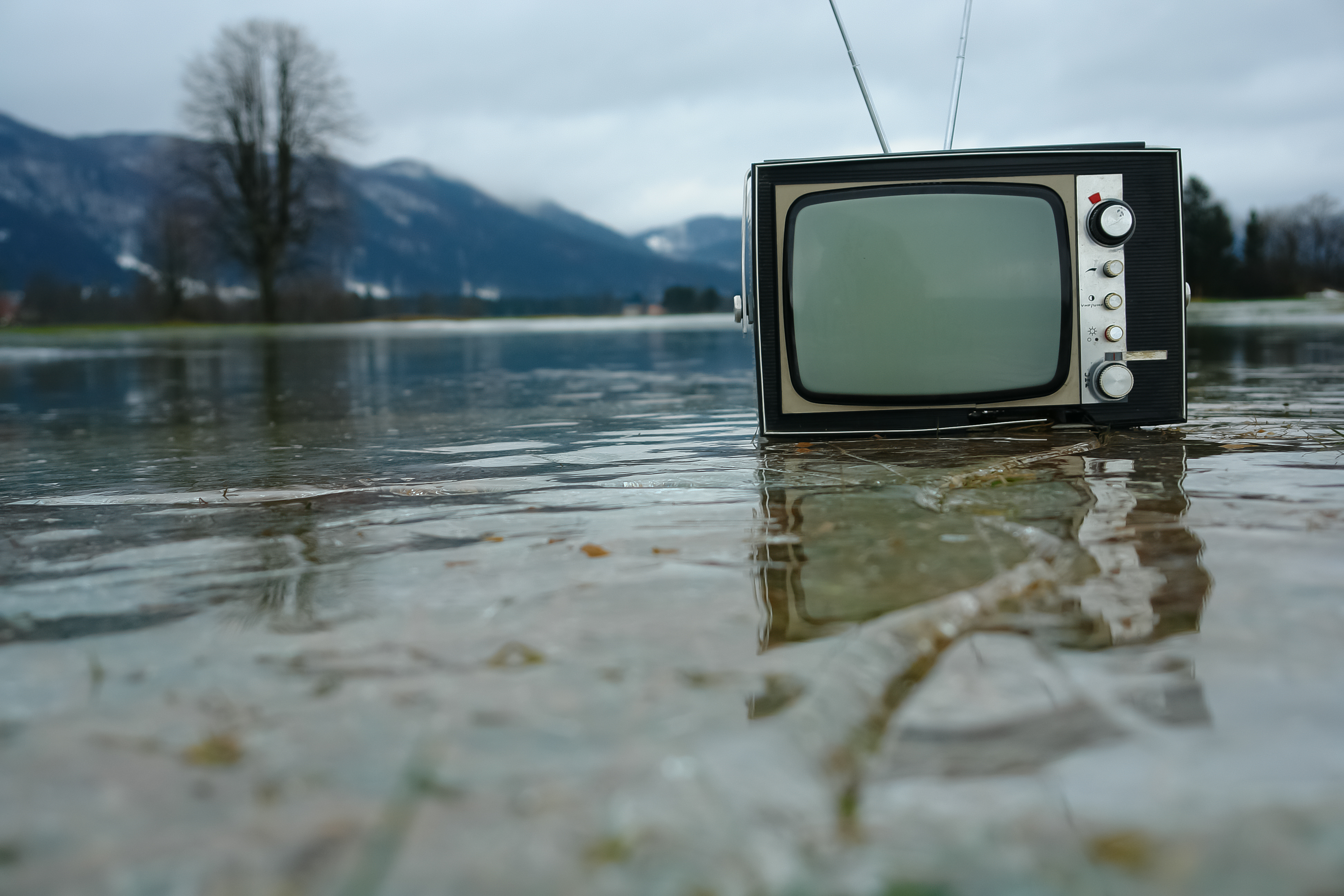 Ein Fernseher steht in einem flachem Gewässer, im Hintergrund eine Landschaft mit Bäumen und Bergen. 