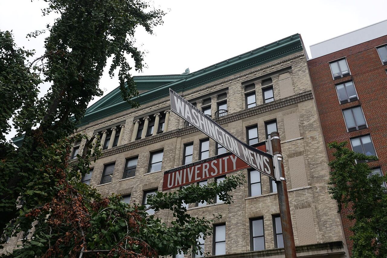 A corner street sign in fron of a large, elegant, white brickstone building. The signs read: University Rd. and Washington Mews