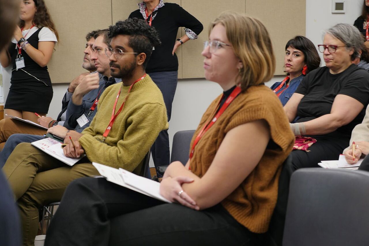 Close up shot of the audience. Two rows of people are looking to the front, two people standing in the background.