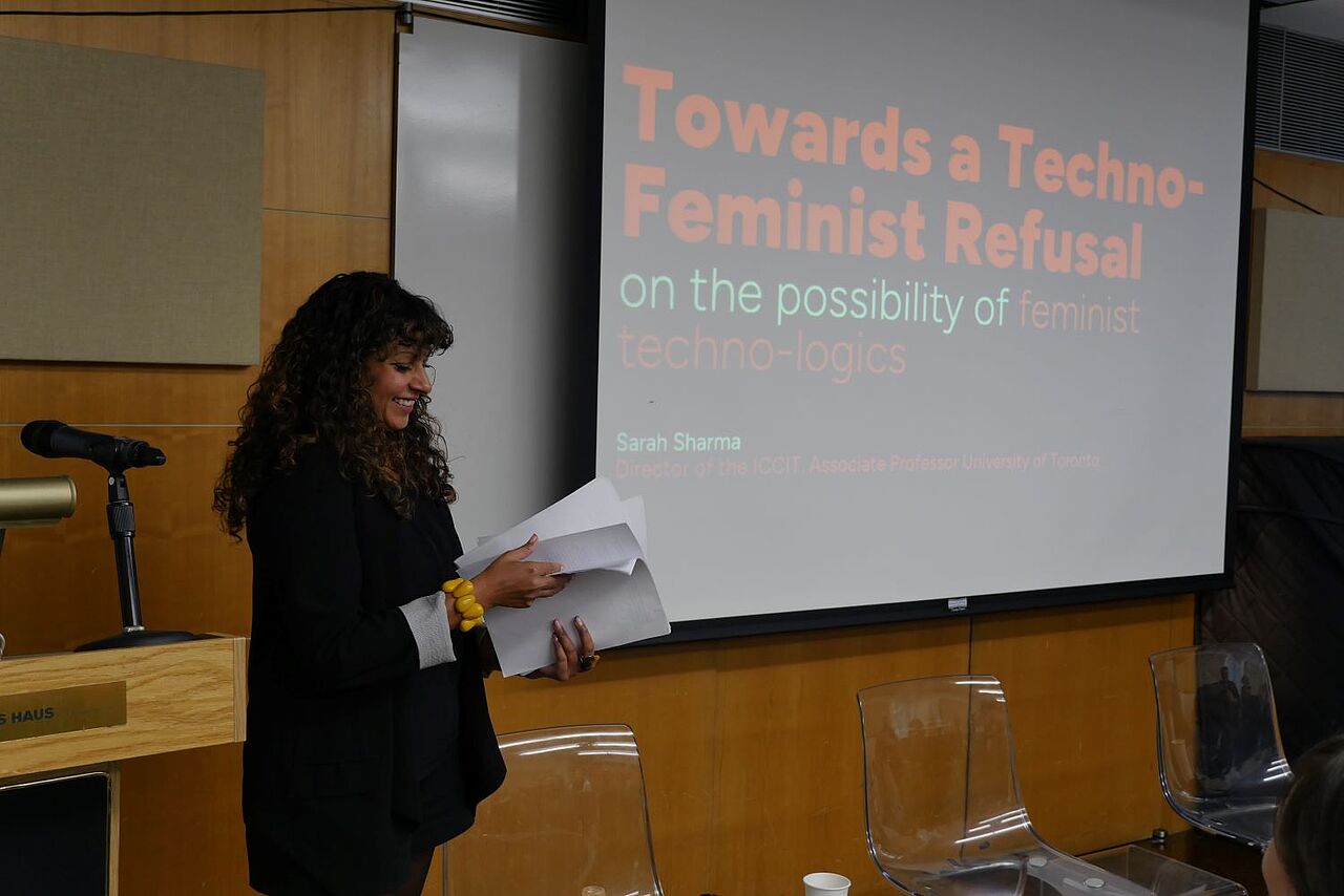 Profile of Sarah Sharma standing in front of the podium, sheets of paper in her hand, smiling. The slide behind her reads: Towards a Techno-Feminist Refusal. On the possibility of feminist techno-logics.