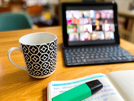 Das Bild zeigt einen Tisch mit einem Kaffeebecher, einem Notizbuch sowie einem Textmarker. Verschwommen im Hintergrund ist ein Monitor zu sehen, auf dem aktuell eine Videokonferenz stattfindet. 