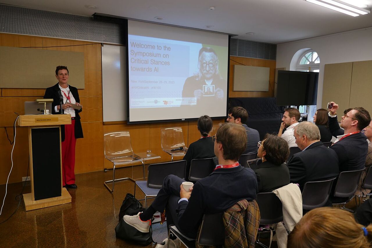 Ricarda Opitz standing in front of the audience, giving her opening remarks. The slide behind her shows Joseph Weizenbaum and "Welcome to the symposium on critical stances towards AI". 