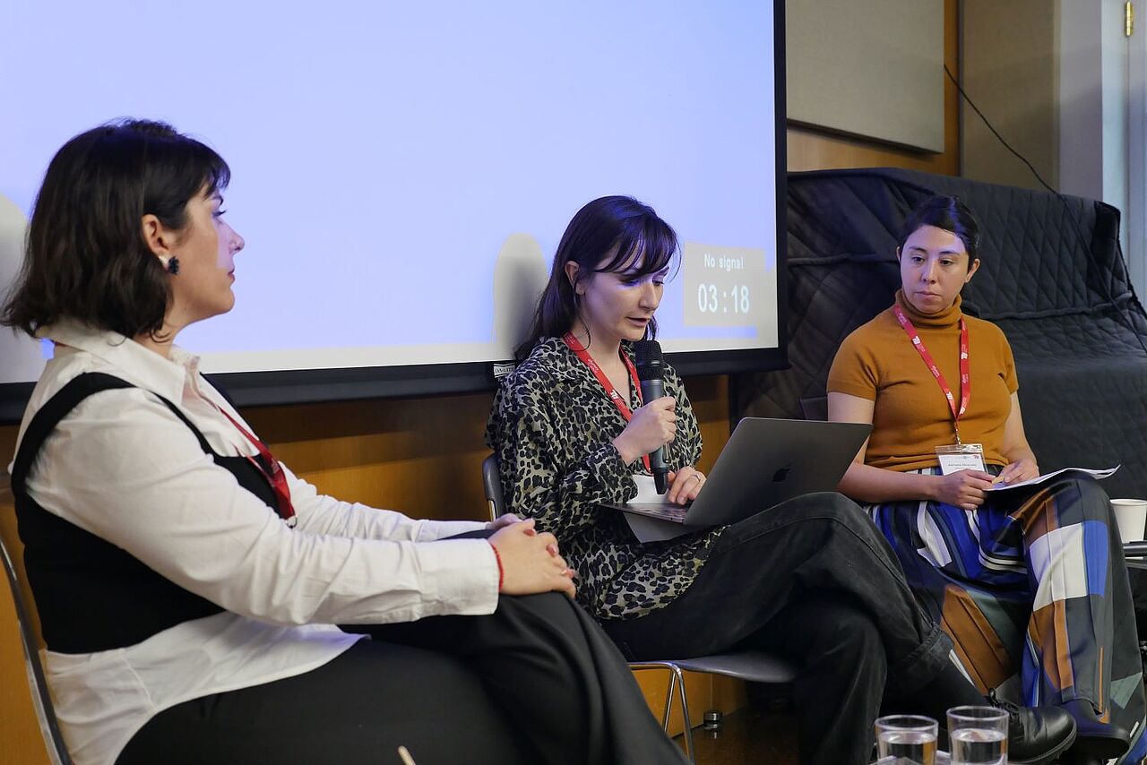Three women sitting on chairs next to each other. Milagros Miceli in a black and white shirt. Alexandra Keiner in the middle with a microphone in hand and a laptop on her lap. Adriana Alvarado with a yellow shirt on the right. 