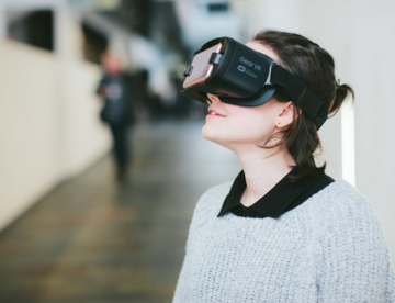 Eine weiße Frau mit braunen Haaren und hellem Pullover trägt eine VR-Brille und schaut leicht nach oben. 