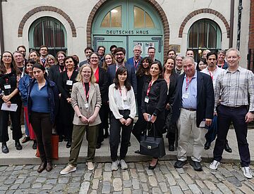 A group of ca. 30 people standing on a street, in front of the green door, the entrance Deutsches Haus at NYU. 